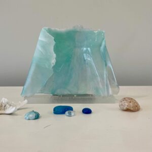 A translucent blue-green resin art piece resembling sea waves, accompanied by a small seashell, coral, and three polished stones on a table.