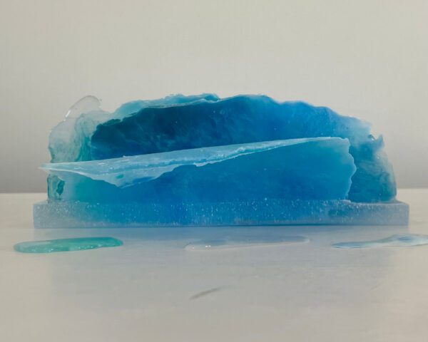 A translucent blue resin sculpture resembling an ice formation, displayed on a white surface with droplets around it.