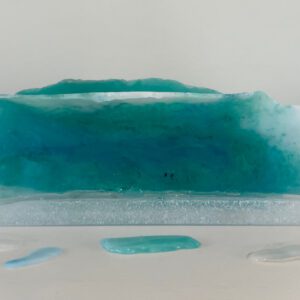 A translucent sculpture resembling a wave with gradients of blue and green, displayed on a white surface with water droplets around it.