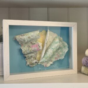 A framed piece of Peggy Paisley cloth on a shelf, flanked by a white jar and stacked towels.