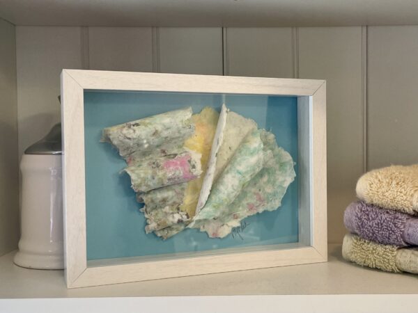 A framed piece of Peggy Paisley cloth on a shelf, flanked by a white jar and stacked towels.