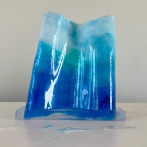 A translucent blue sculpture reminiscent of a frozen waterfall, displayed on a white surface with small scattered shards.