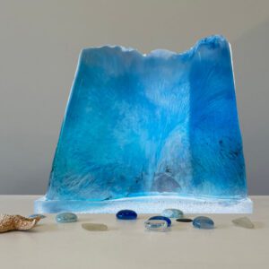 A translucent blue resin sculpture resembling a wave, displayed with scattered glass pebbles and a seashell on a light surface.