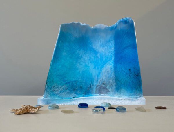 A translucent blue resin sculpture resembling a wave, displayed with scattered glass pebbles and a seashell on a light surface.