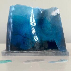 A blue resin sculpture resembling a wave, displayed on a gray base with small puddles nearby, set against a plain background.