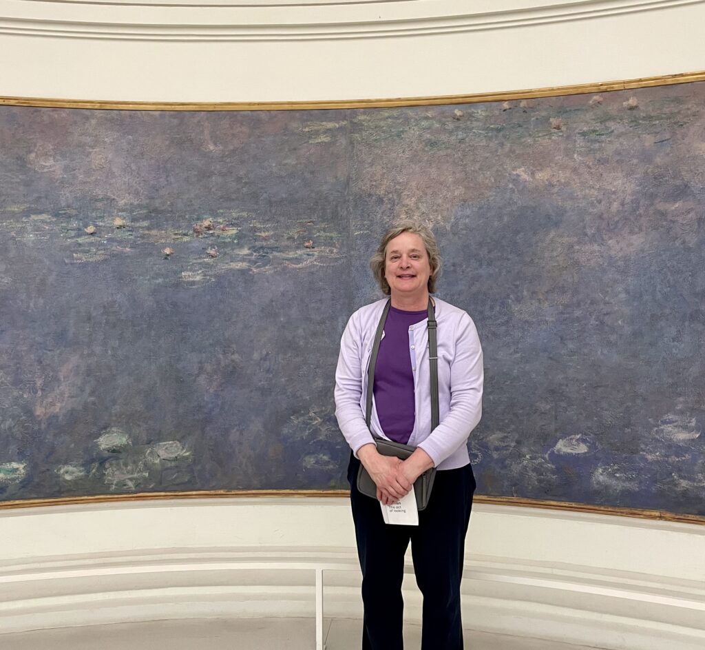 A woman stands smiling in front of claude monet's "water lilies" painting in a gallery.