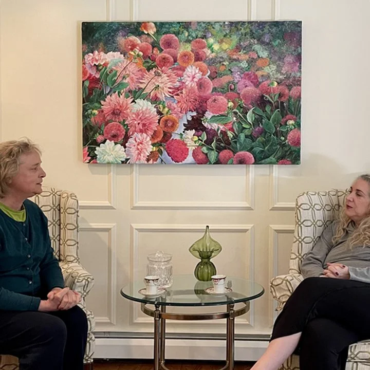 Two women sitting in chairs with a floral painting.